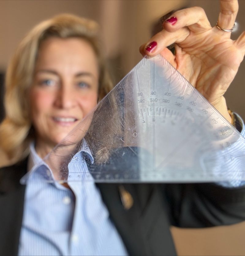 En kvinde (Anna Foss) holder en gennemsigtig vinkelmåler foran sig. Vinkelmåleren er i fokus, hvilket gør kvindens ansigt sløret. Hun smiler og ser ud til at pege på en af vinkelmålerens markeringer. Kvindens negle er malet røde, og hun bærer en lyseblå skjorte samt en sort blazer. Medietræning budskabsudvikling vinkel kernebudskab kommunikation